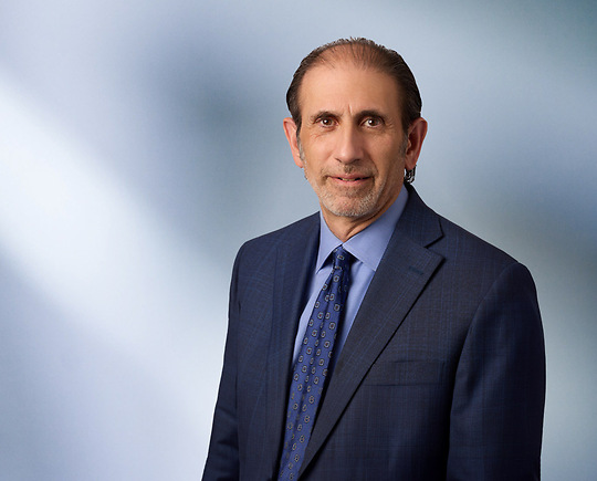 A man, Jonathan David, in a dark blue suit, light blue shirt, and patterned tie is looking at the camera, standing against a blurred blue and white background.