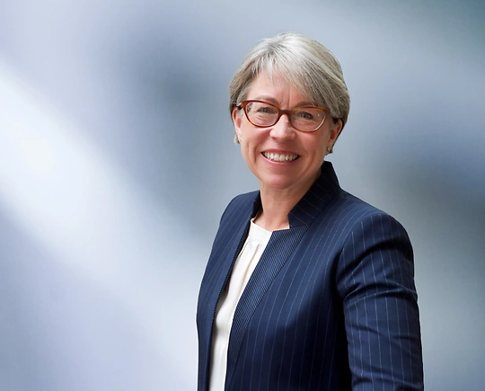 A woman, Deanna Swisher, in a navy pinstripe suit, white blouse, and glasses is smiling at the camera, standing against a blurred blue and white background.